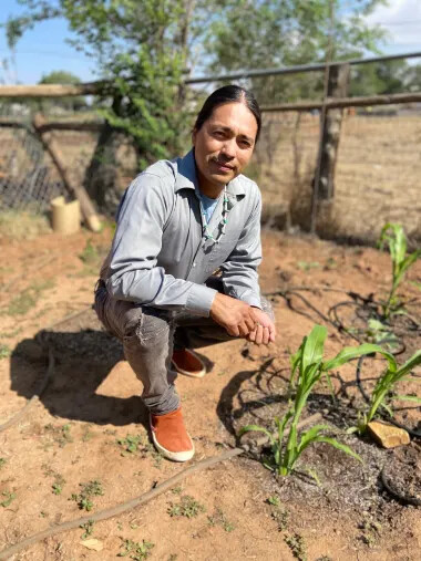 A black-haired man with a blue button down shirt, gray jeans, and orange shoes squats low towards sandy ground above the green shoots of young plants. 