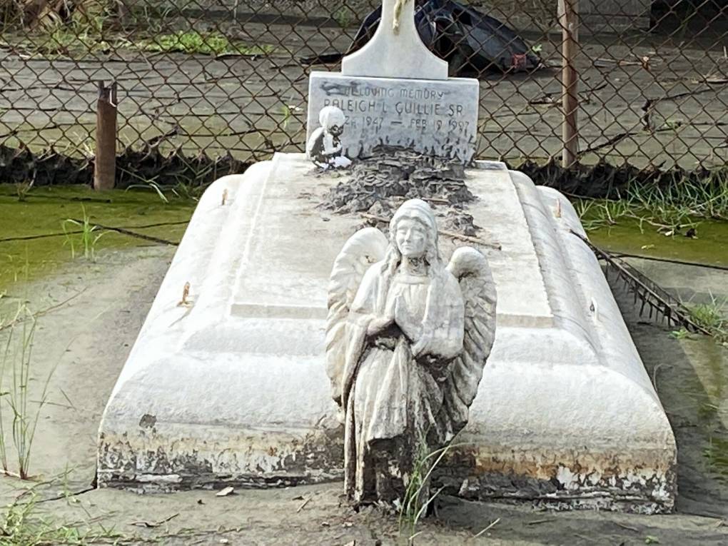 A white stone angel covered in black moss sits in front of a grave site covered in mud.