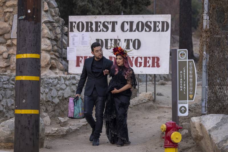 A man and pregnant woman wearing dressy black clothing are walking near a sign in the Angeles National Forest that reads Forest Closed Due to Fire Danger. The couple were on an outing to pose for pregnancy photos.