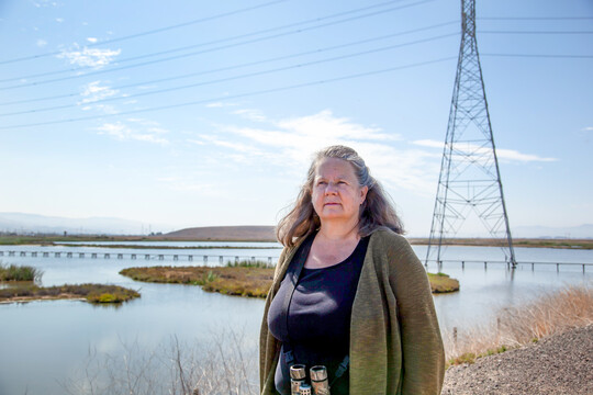 A woman with gray, shoulder-length hair wears a green sweater and silver binoculars around her neck. A wetland and powerlines are in the background against blue skies. 