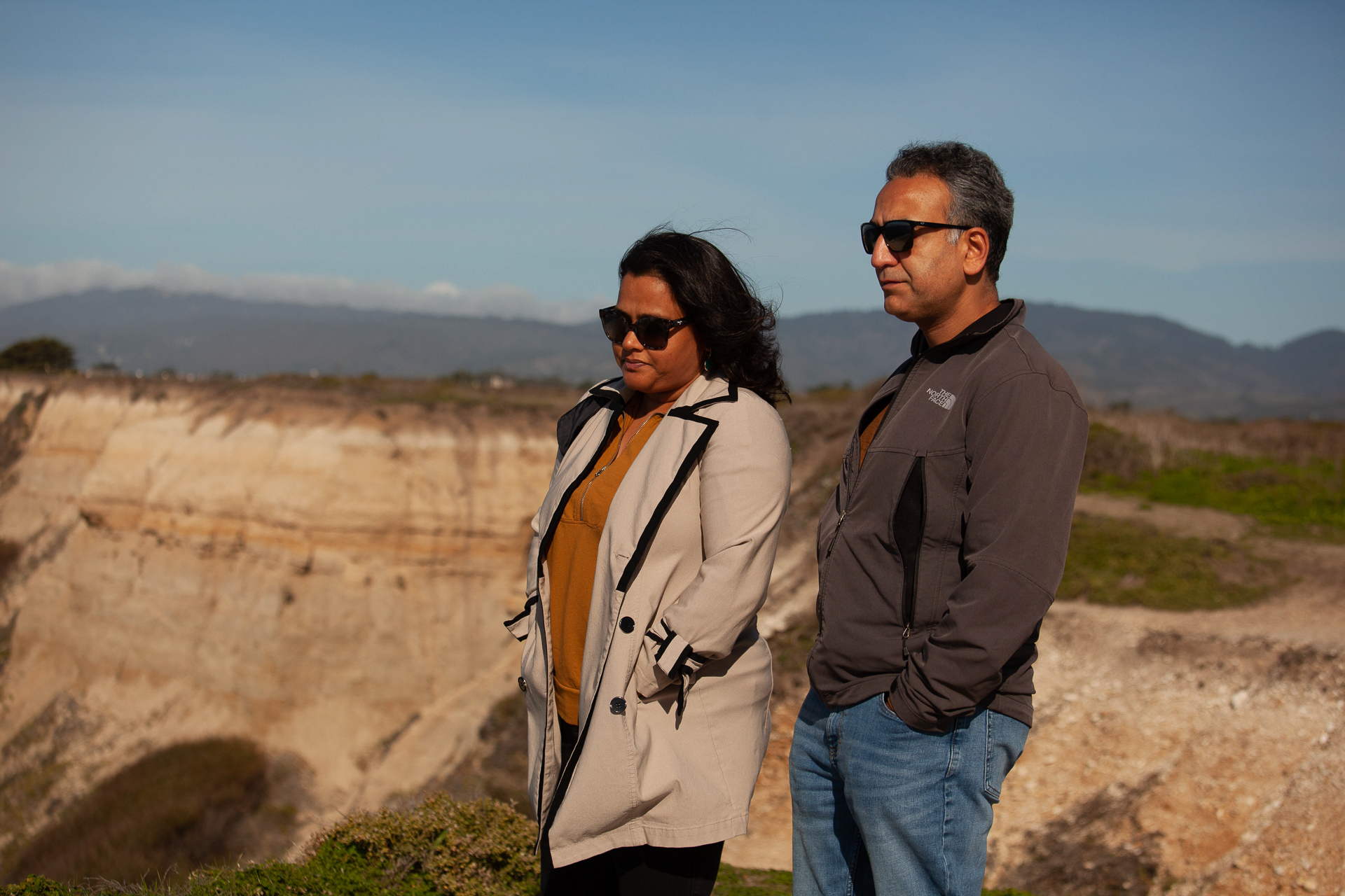 A women with a beige trenchcoat and a man with a gray fleece stand on a bluff looking over the ocean. 