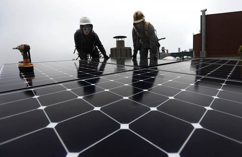 A solar panel is installed by two workers on a rooftop. The workers are wearing hard hats and are at the far background of the frame.