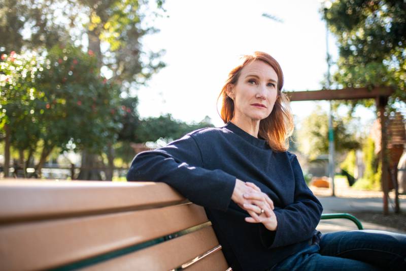 A woman with red hair sits at the end of a brown bench, a playground and sunshine in the background.