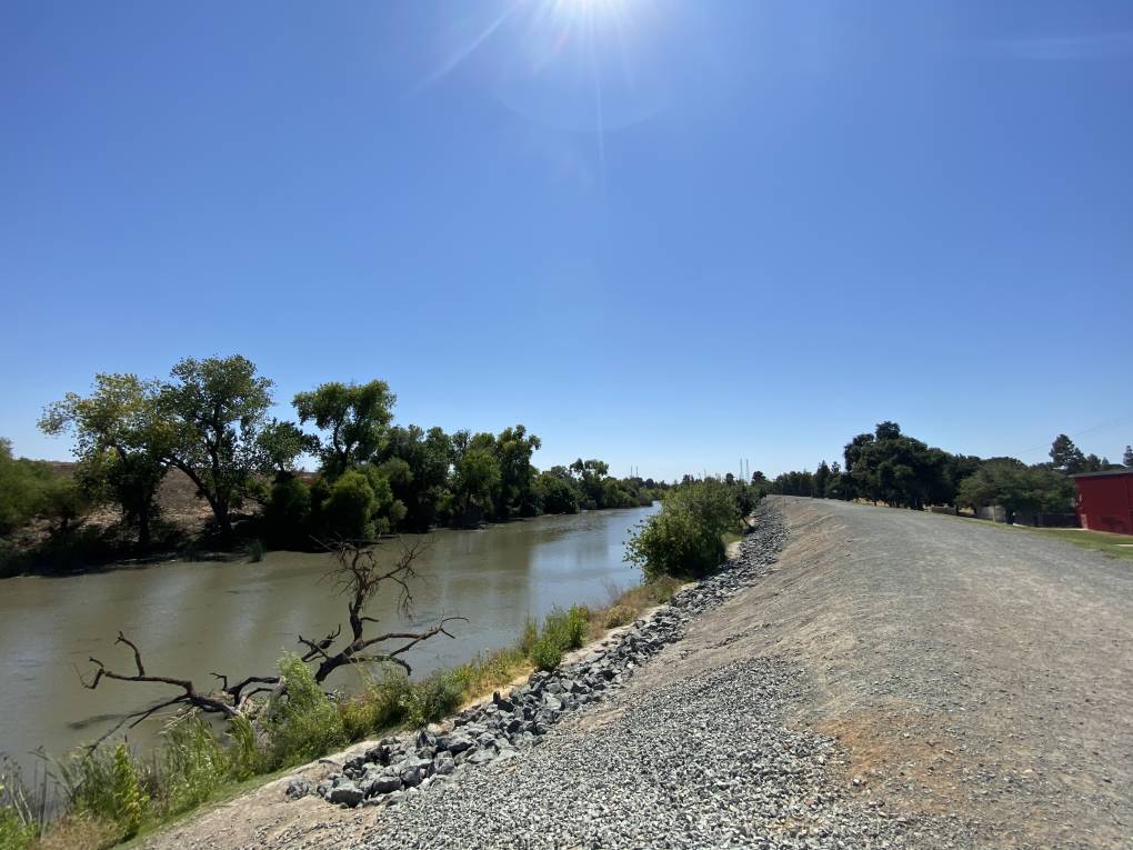  A view down a long, earthen, rocky levee that runs alongside a green river about the same width, with scrabbly green trees on all verges, beneath a clear, sunny, blue sky.