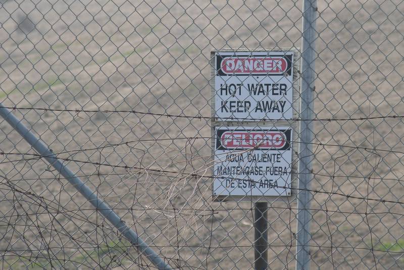 A sign is posted behind a chain link fence, protected with strands of barbed wire. The sign, in Spanish and English, reads "Danger. Hot Water. Keep Away."