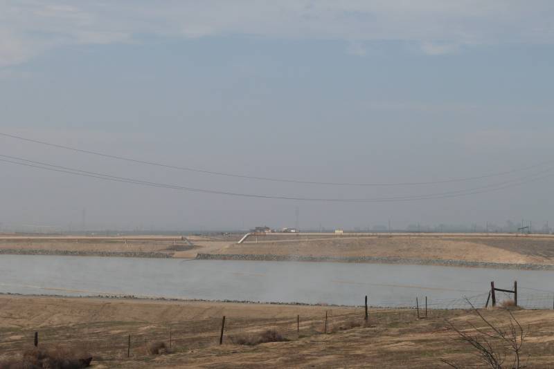 White steam rises off gray water at a Cawelo Water District reservoir filled with wastewater from Chevron's Kern River Oil Field. In the foreground and behind the oval reservoir are brown fields. A long pipeline extends from the far right of the photo to the edge of the reservoir.