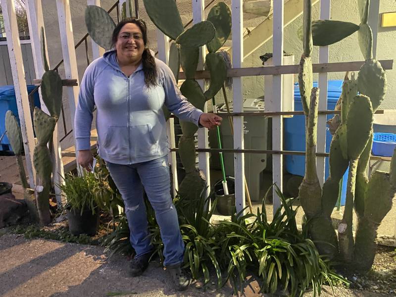 A smiling Mexican woman wearing blue jeans and a light blue hoodie stands smiling in front of the two story apartment building where she lives. Tall cactus plants hug the fence behind her.