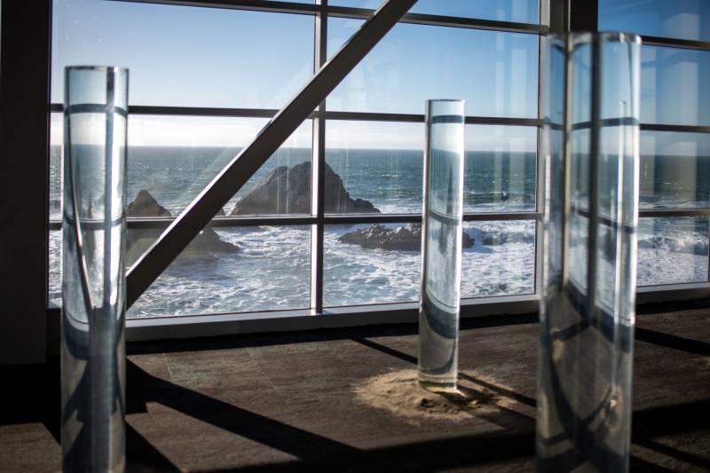 Three glass cylinders, or sea bodies, six feet tall, rise from a pile of sand in front of wide glass windows at San Francisco's Cliff House. The sea green ocean beyond the windows is restless with whitecaps. Dark mounds of rock rise from the sea.