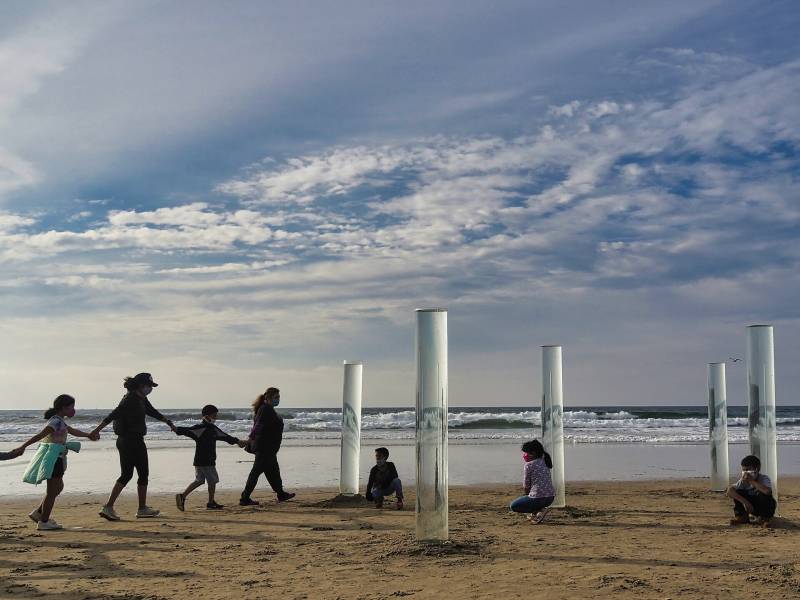 Five clear, cylindrical pillars of ocean water emerge vertically from the sand on a shoreline. Three children sit at the bases of the scattered pillars, while four people hold hands and walk towards them from the left of the frame.