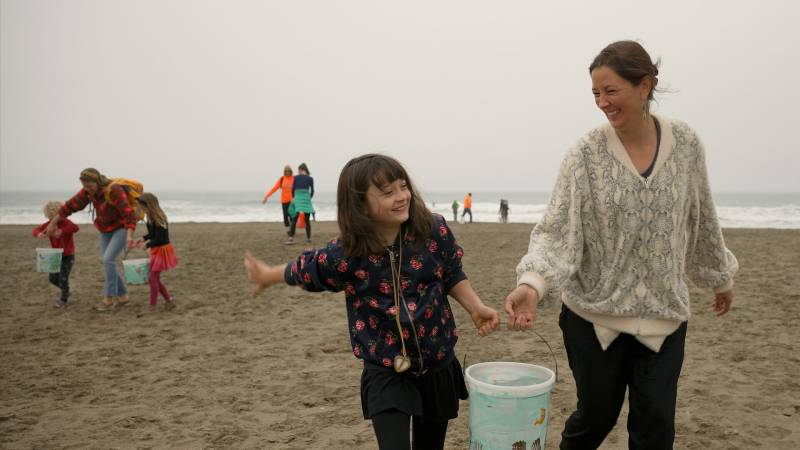 A mother and child smile while carrying a white bucket painted light blue between them. They walk across a beach toward the camera and away from the ocean. More people carrying buckets follow behind.