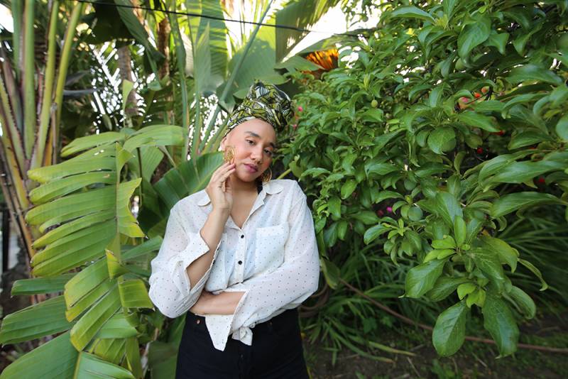 Woman stands in front of lush foliage, looks seriously into camera. She’s wearing a white shirt with black polka-dots and a green and white head scarf.