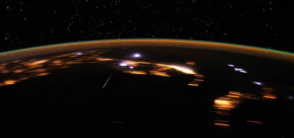 A golden streak arches across the black sky, with tiny dots of stars in the background. In the foreground, blurred streaks of gold flash through the black space in lines and ovals. This is an image of a Lyrid meteor caught on video camera from aboard the International Space Station.