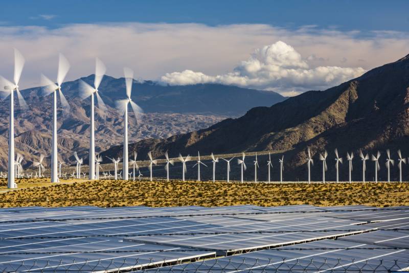 Glistening rows of blue solar panels line the desert floor in the foreground. Beyond them are rows of spinning white windmills, set against a backdrop of craggy brown mountains. In the distance a shadowed black mountain shrugs across the horizon. Puffy white clouds nestle into the mountain with streaks of thin white clouds behind and a blue sky.