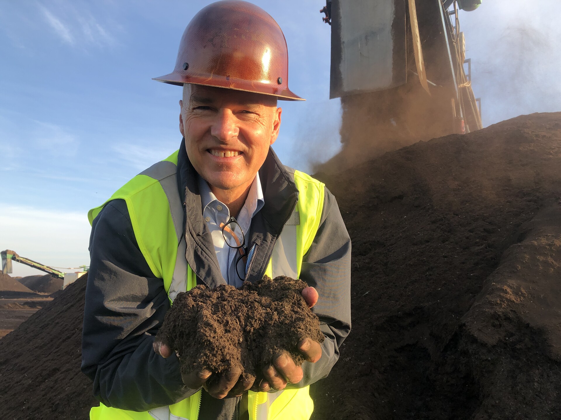 A smiling man with a red hard hat holds a handful of rich, brown compost as a crane dumps more in a large pile behind him.