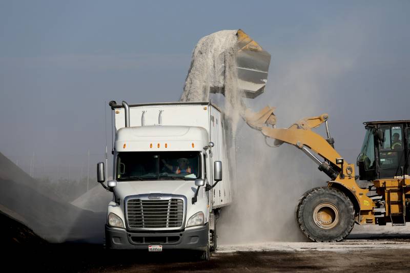 A frontal view of a white semi, with a yellow bulldozer beside it dumping a load of white compost into its trailer, which is sending up a cloud of compost dust. The sky is blue behind them, with low, dark gray hills of maybe dirt of their other side.