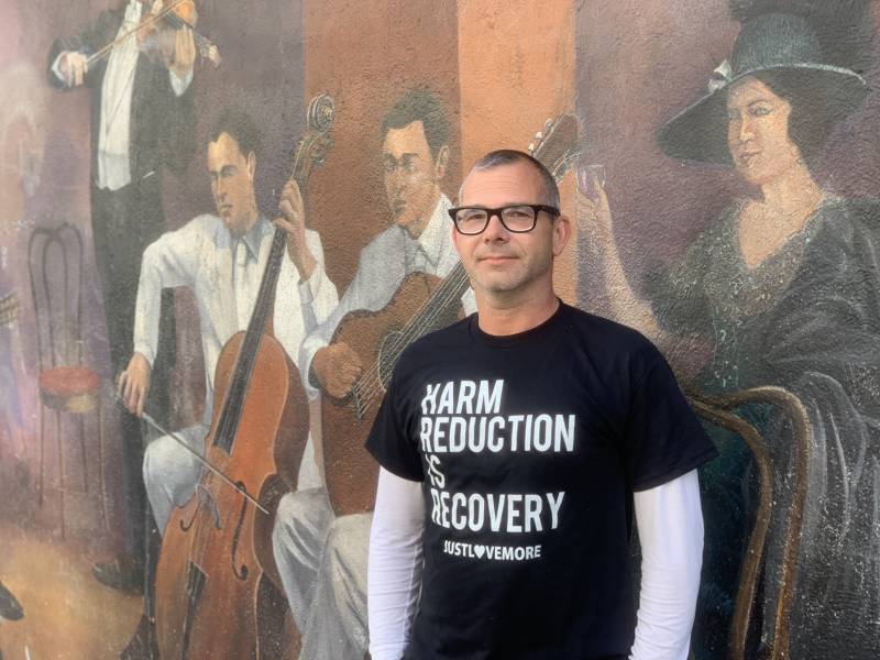 A man with white skin stands in front of a huge wall mural showing musicians. The man has a very short buzz cut and wears black glasses, and a long-sleeved white t-shirt under a short-sleeved black t-shirt that reads, "Harm reduction is recovery." The three male musicians in the mural are dressed in formal wear of all white or a tuxedo. One plays a violin, one a base, and one a guitar. A woman on the right side of th emural is dressed in a formal gown of black with white sequins, a lush dark brown cape and a black hat with a wide brim.
