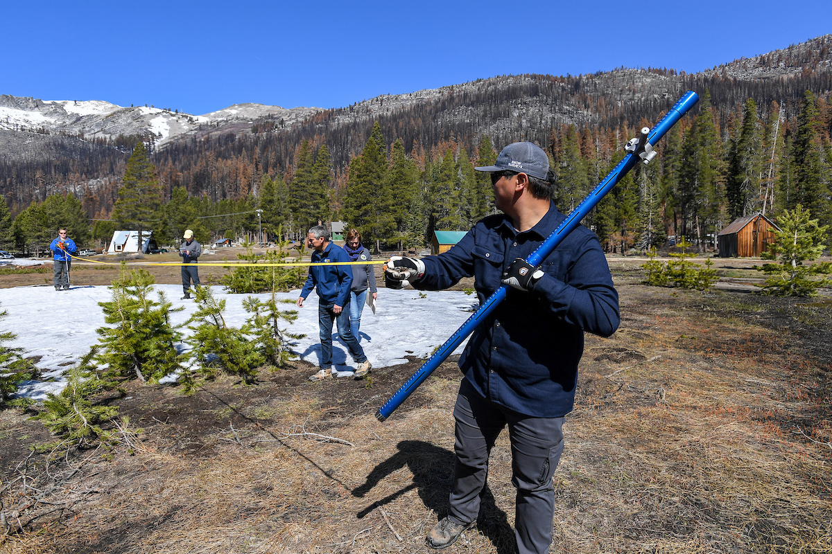 Man holds a hollow blue pole that when pierces the ground tells how much water is in snow in a certain spot. Blue skies and green mountains in the distance. A patch of snow in view. 