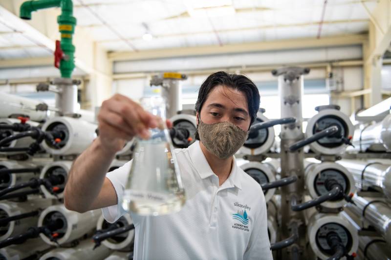 An Asian man in a white polo shirt holds a beaker full of water that's been filtered. A system of tubes behind him.