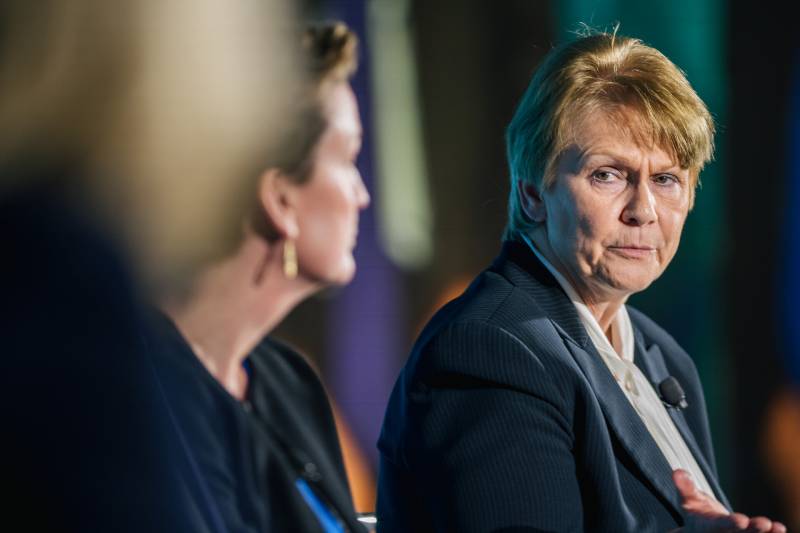 An older woman in a navy blue suit jacket and white shirt is at the far right of the photo. She has dark blond hair and is looking a woman in a blurred foreground to the left. The woman featured in the photograph is the CEO of Occidental Petroleum, Vicki Hollub.
