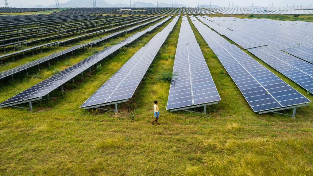 A dark-chocolate skinned man wearing blue shorts and a yellow shirt walks past a gleaming array of solar panels that fills a vast field of yellow-green grass, far out to the horizon. The photo is a solar farm in Karnataka, India.