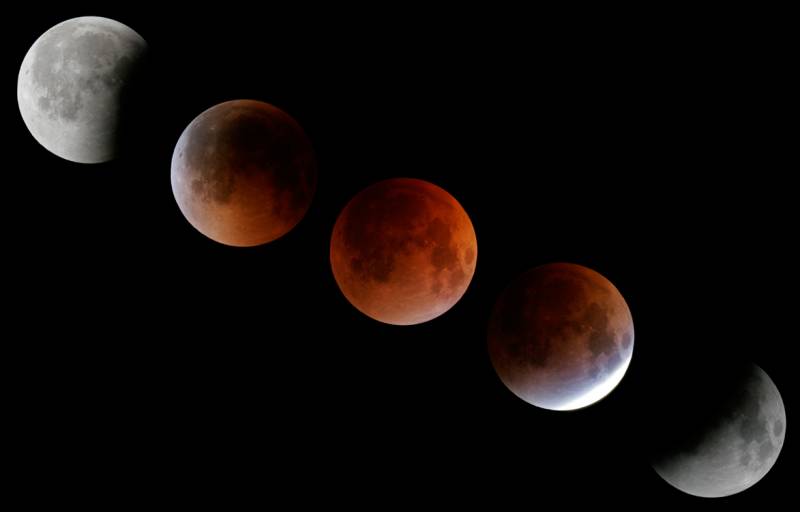 Five moons move from lower right to upper left, showing the progression of a total lunar eclipse. The first moon is one-half dark gray, with a black bite taken out of the left side, as the moon moves toward Earth's shadow. In the second phase, there's a sliver of bright sunlight on the lower right side of the moon, while most of the moon is dark with a slightly reddish cast. In the third phase, the moon is fully in Earth's shadow and covered in a red/orange light. In the fourth phase, the moon is moving out of Earth's shadow and is dark grey with a reddish cast and pale grey on the right side. In the fifth and final phase, there's a dark shadow on the right quarter of the moon, while the rest is white and grey.
