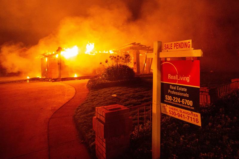 A photo taken at night that is mostly orange. A real estate sign hangs on a white post in front of the lush lawn of a burning home. The sign says "Sale Pending." In the background, a house is encased in fire, with bright flames and smoke, lit orange by the fire, billowing all around it.