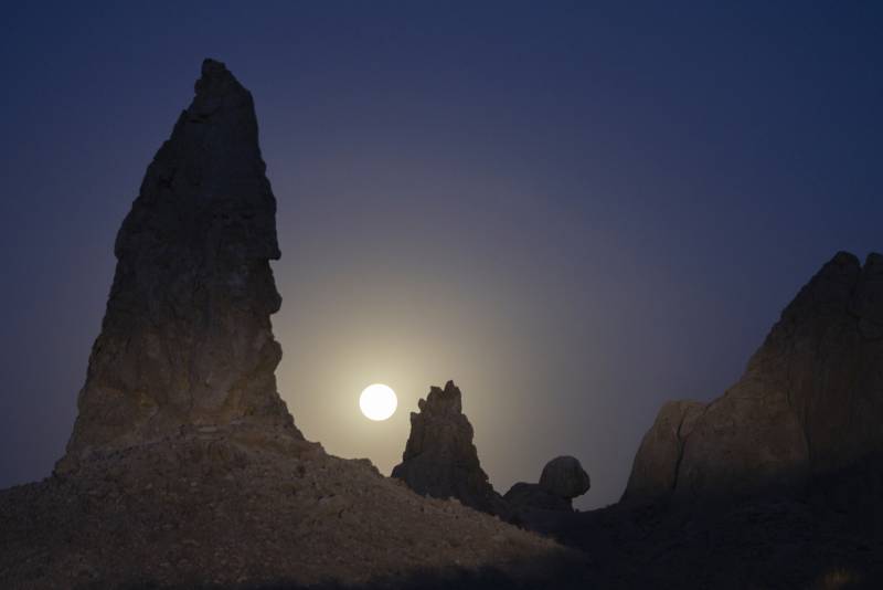 A large bright, white moon glows in the sky, hovering between two dark and craggy rock formations shaped like towers. The sky is blue-black.
