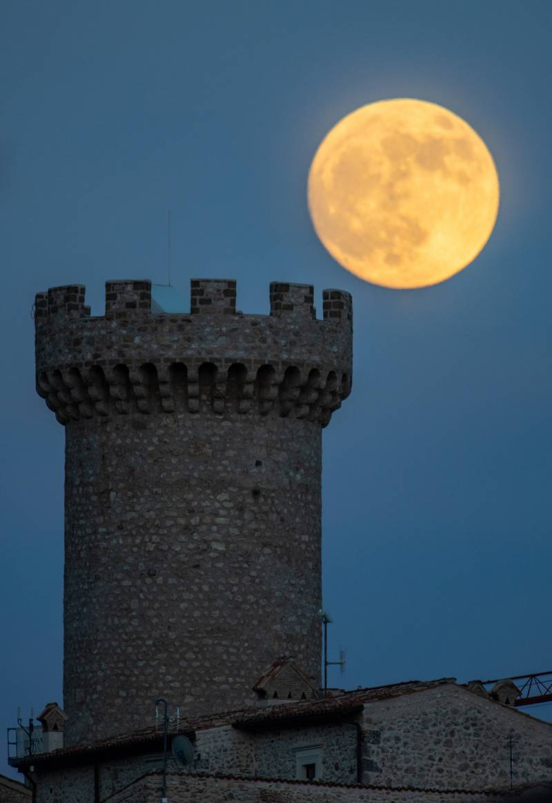 A huge full moon glowing yellow and orange looms over a medieval tower, a stone column with turrets at the top. Behind is a dark blue sky.