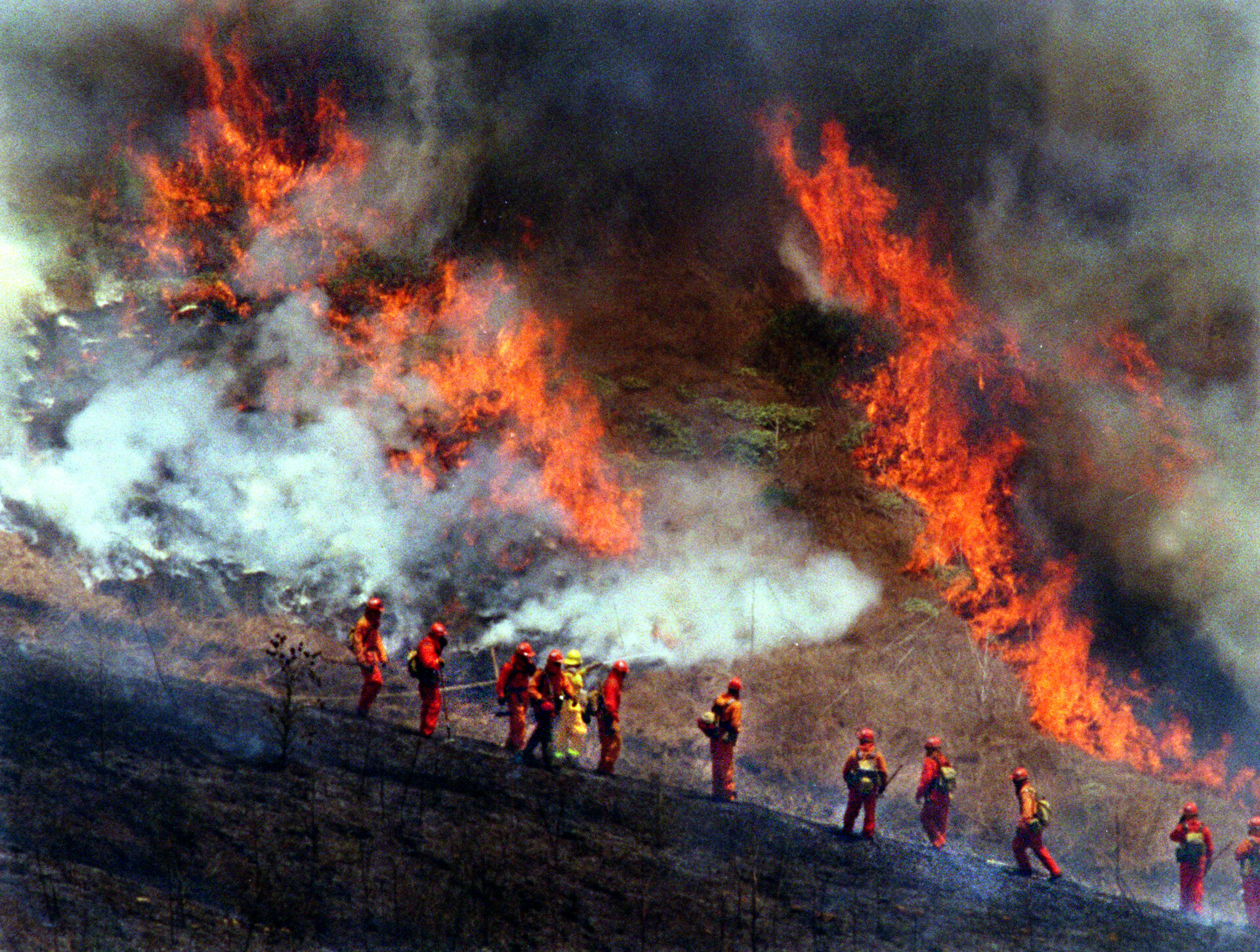 Firefighters in orange and yellow uniforms stand in front of a burning hillside.
