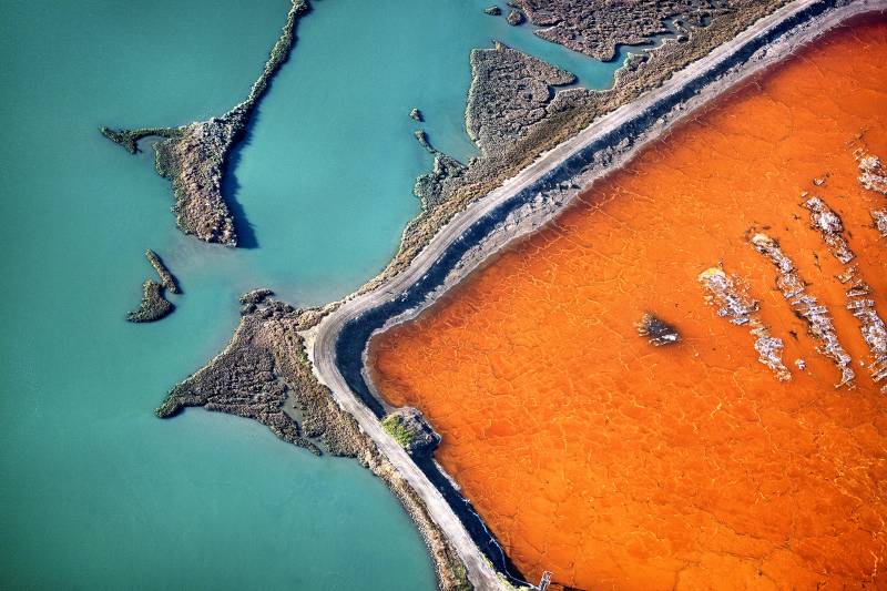 A bright orange square intrudes from the lower right corner of the image, bumping up against a levee of white and blue, separating the salt pond from the bay. Landscapes of green and brown spill in fractal shapes from the levee into the a pale turquoise water of the bay.