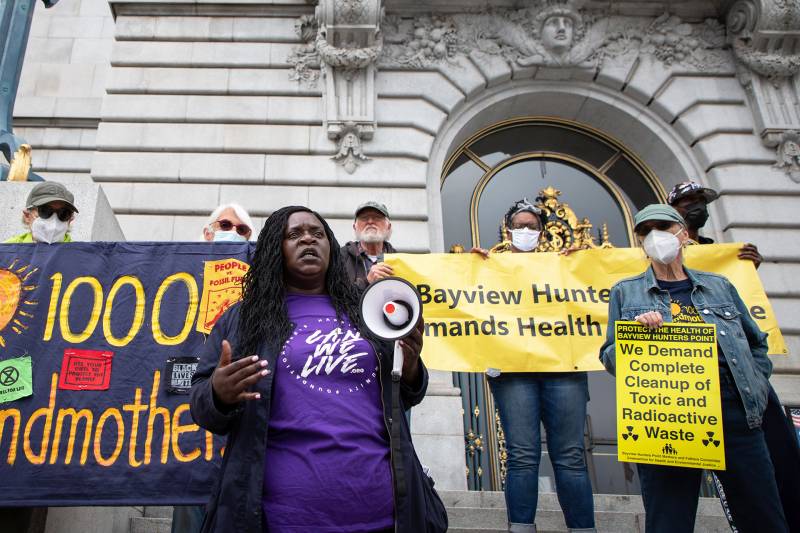 A Black woman with long black braids is wearing a purple shirt that reads 'Can we Live.' People are standing on the cement steps of San Francisco City Hall holding signs about environmental justice for the neighborhood of Bayview-Hunters Point.