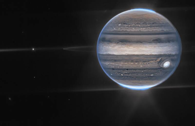 A wide field view showcases Jupiter in the upper right quadrant. The planet’s swirling horizontal stripes are rendered in blues, browns, and cream. Electric blue auroras glow above Jupiter’s north and south poles. A white glow emanates out from the auroras. Along the planet’s equator, rings glow in a faint white. These rings are one million times fainter than the planet itself! At the far left edge of the rings, a moon appears as a tiny white dot. Slightly further to the left, another moon glows with tiny white diffraction spikes. The rest of the image is the blackness of space, with faintly glowing white galaxies in the distance.