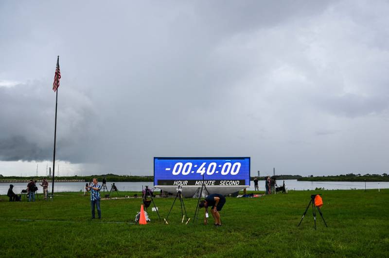A cloudy sky with a large, bright blue multipanel countdown clock set at 00:-40:00 (hours, minutes, seconds). In the foreground a couple of men appear to be packing up camera tripods on a grassy lawn.