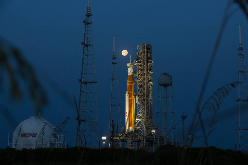 The image shows a full moon glowing over the tip of a spacecraft, set up on a launchpad. Other iron towers rise into an indigo sky. A blurred branch drifts in from the left of the image, in the foreground.