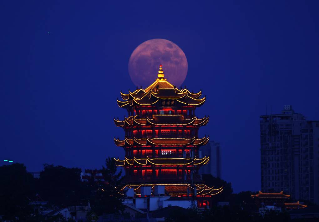 Yellow light outlines the pagoda shapes of a tall tower against a vivid blue sky. Behind the tower is a huge supermoon in pink and purple.
