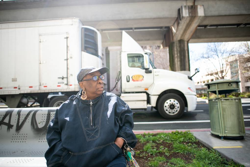 A black woman with a blue jacket, baseball hat and long ear sits on a bench. A white semi-truck passes behind.