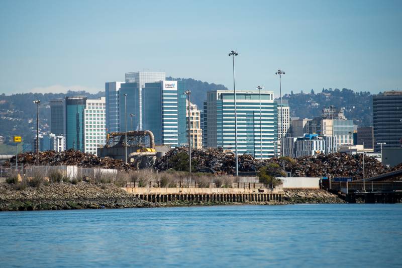 A view from the water of high-rise buildings beneath a pale blue sky. Along the waterfront are piles of scrap metal and industrial pipes. Between the scrap metal and the water is an embankment of rock. In the distant background are wooded hills.