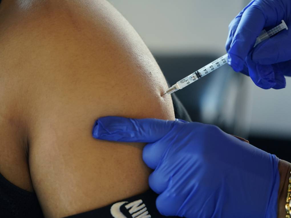 The image shows a closeup of a person's left shoulder and arm. A shot is going into the person's upper arm. One hand in a blue glove is holding the person's shoulder and the other hand in a blue glove is administering the shot.