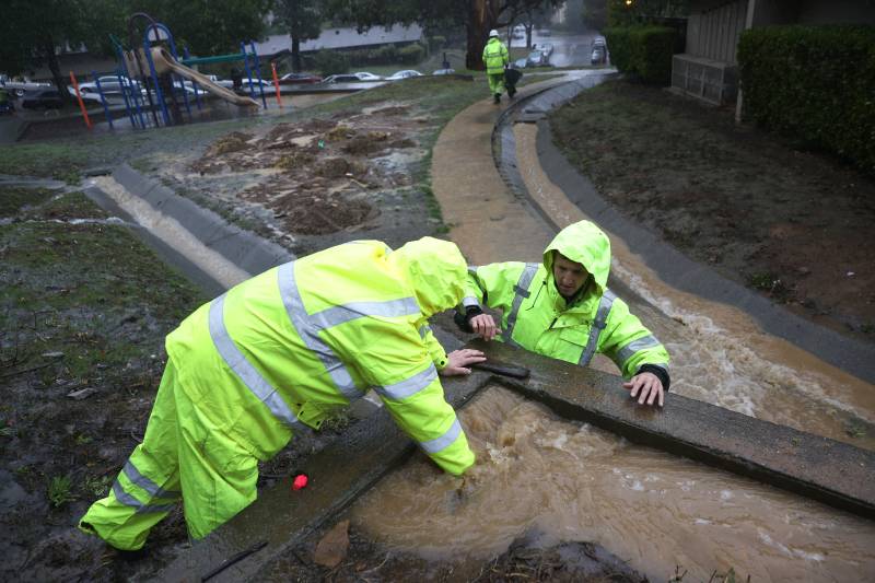 Two people in neon yellow rain gear reach into a flooded storm drain. Behind them is a playground and a stream of water in a grey cement culvert. The water is brown.