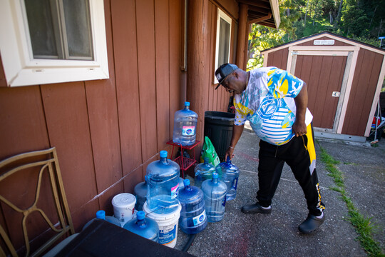 A Black man in a black pants, a white T-shirt and a black baseball cap picks up an empty blue five gallon jug of water. He is near a red outdoor wall of a house and a number of empty five gallon jugs.