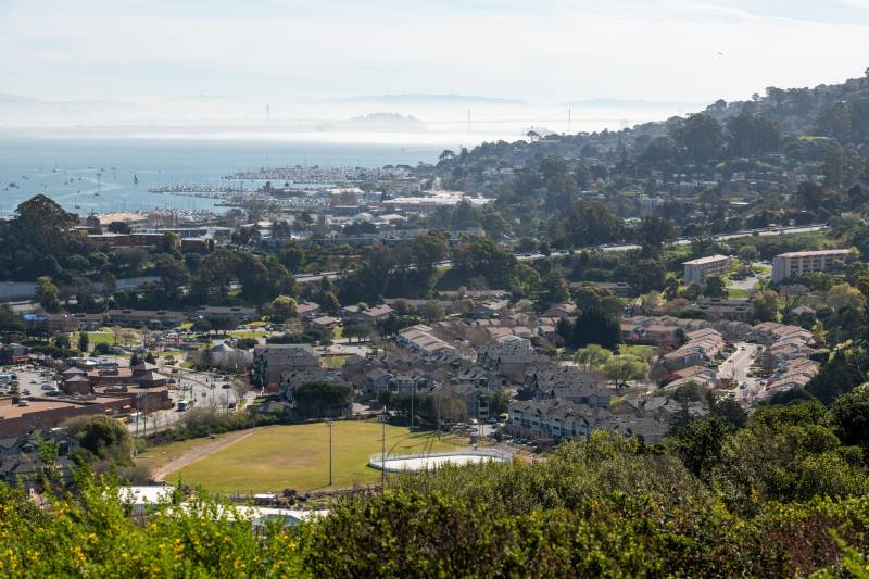 A bright green baseball field in the foreground, surrounded by hills lined with homes and the bay beyond it. Behind the bay there are white clouds and an outline of the San Francisco shore lined with tall buildings.