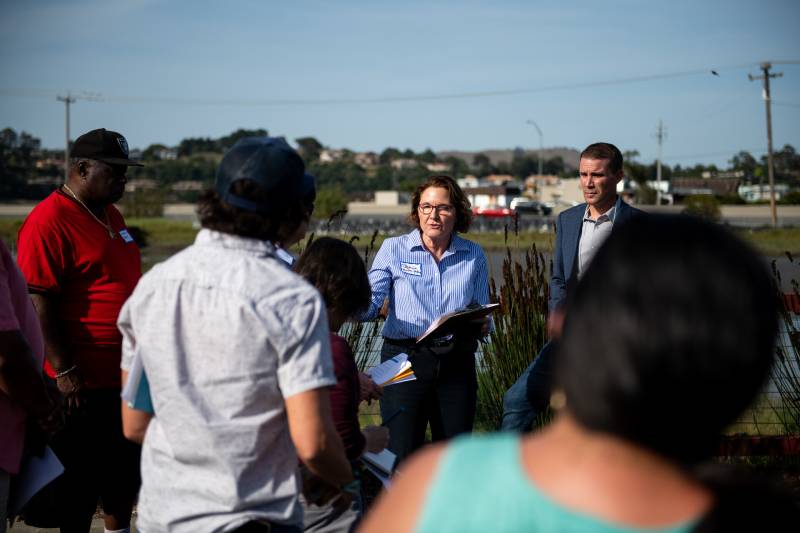 A white woman in a light-blue collared shirt speaks to a crowd of mixed-race people next to a pond.