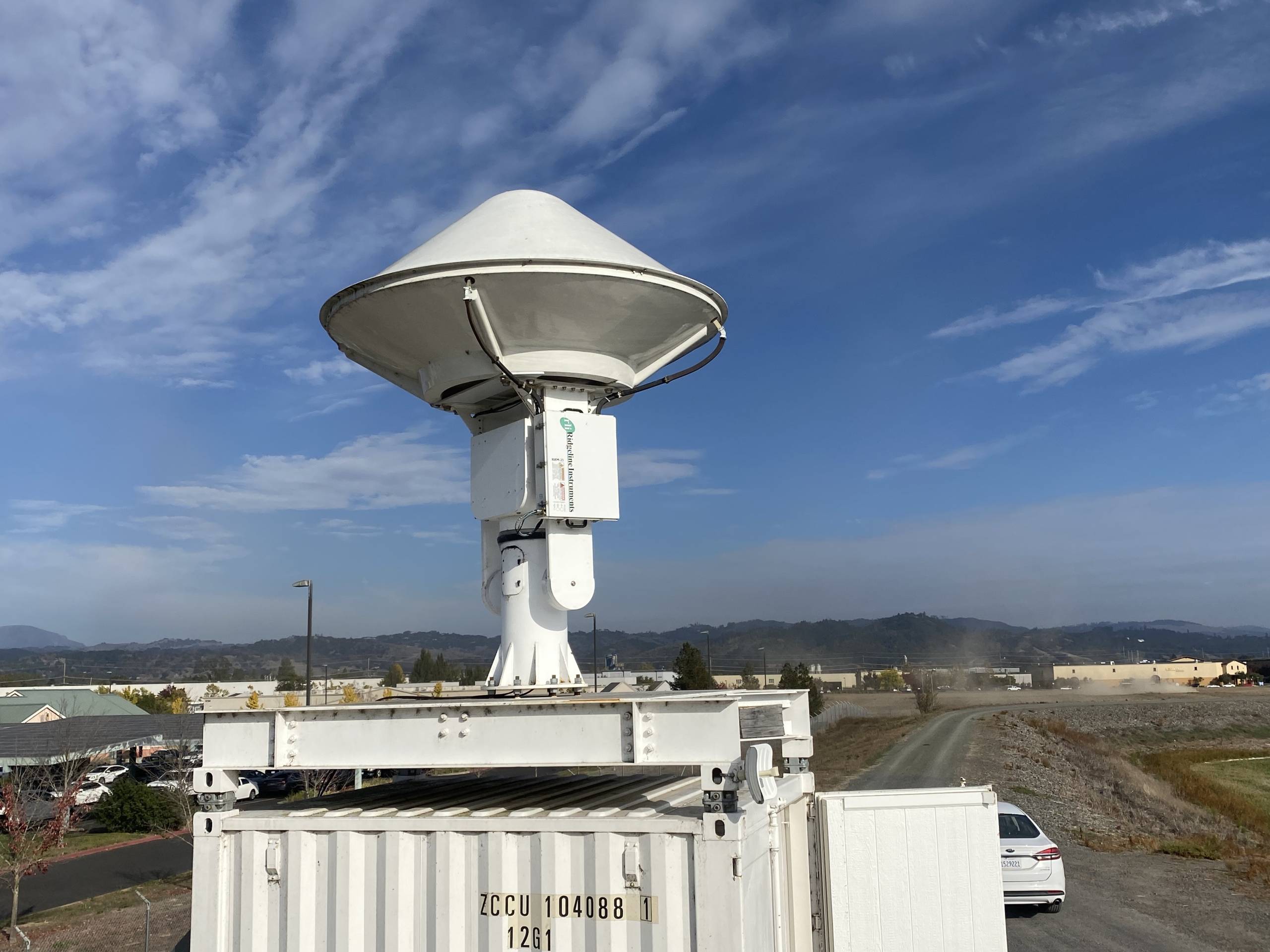 A white radar unit points toward the sky. It looks like a mix between a mushroom and a spaceship. A bright blue sky with wispy clouds is in the background.