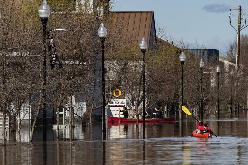 A kayaker in a red boat paddles down a flooded street alongside shops.