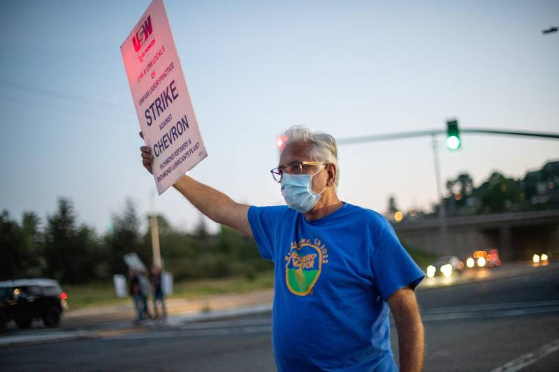 A man wearing a blue shirt with yellow letters holds a white sign reading, 'Strike against Chevron.' A stoplight and fading blue skies are behind him. 
