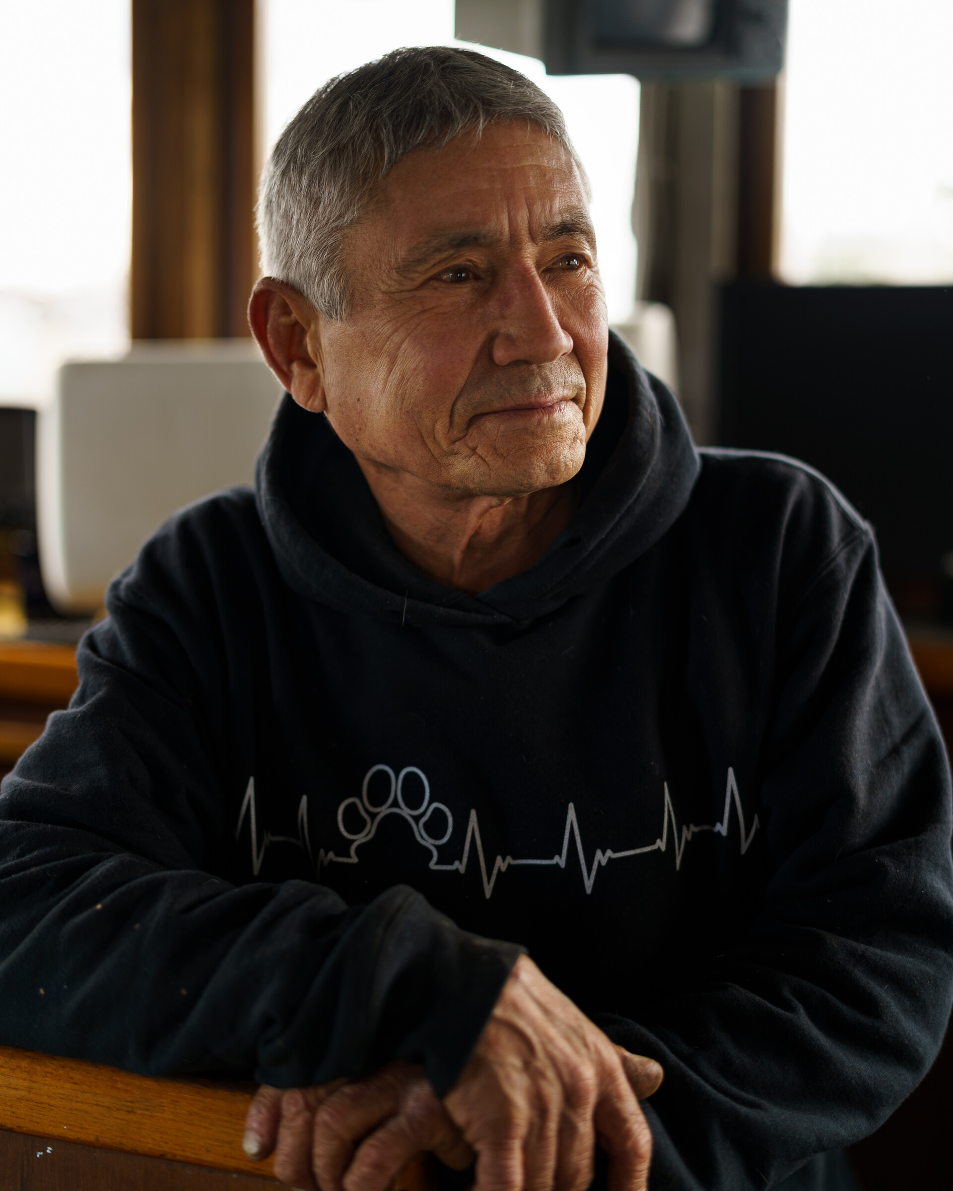 An older man who appears to be of both white and Asian ancestry, with close-cropped gray hair and a black hoodie pictured below-deck of a boat on a bright yet cloudy, glare-y day.