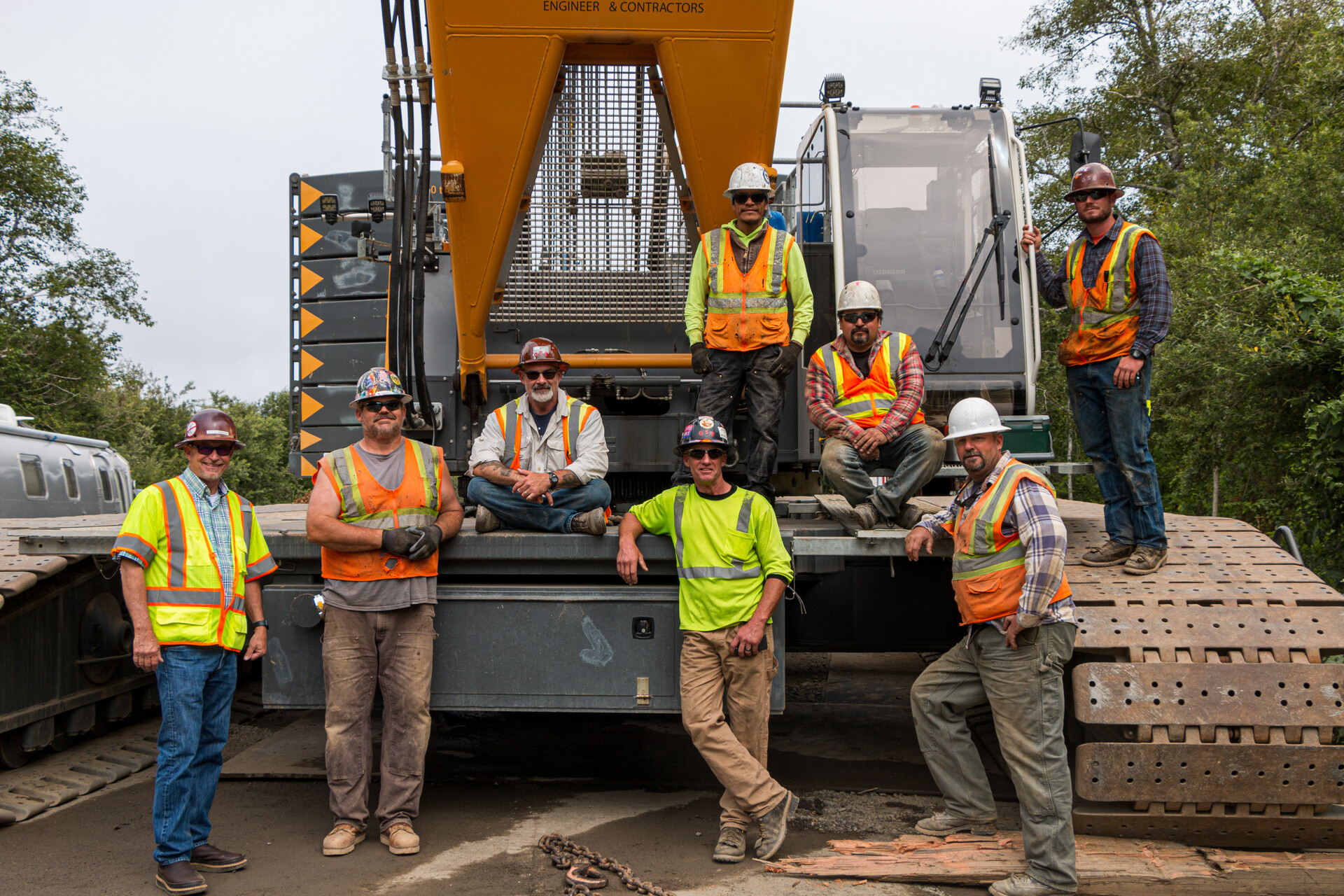 Men in orange, yellow, gray shirts and hard hats stand in front of a crane. 