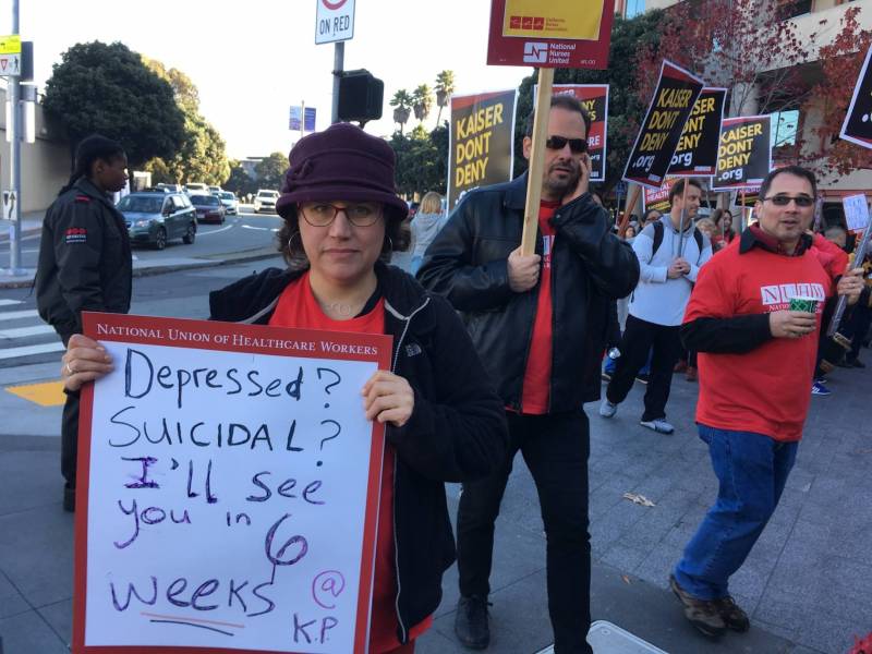A woman with light skin, dark brown hair, glasses, and a purple knit hat holds a sign that says "Depressed? Suicidal? I'll see in you 6 weeks." She faces the camera and stands amid a crowd of people marching on a sidewalk, many of whom are, like the woman, wearing red T-shirts, and holding signs on wooden dowels that say "Kaiser Don't Deny."
