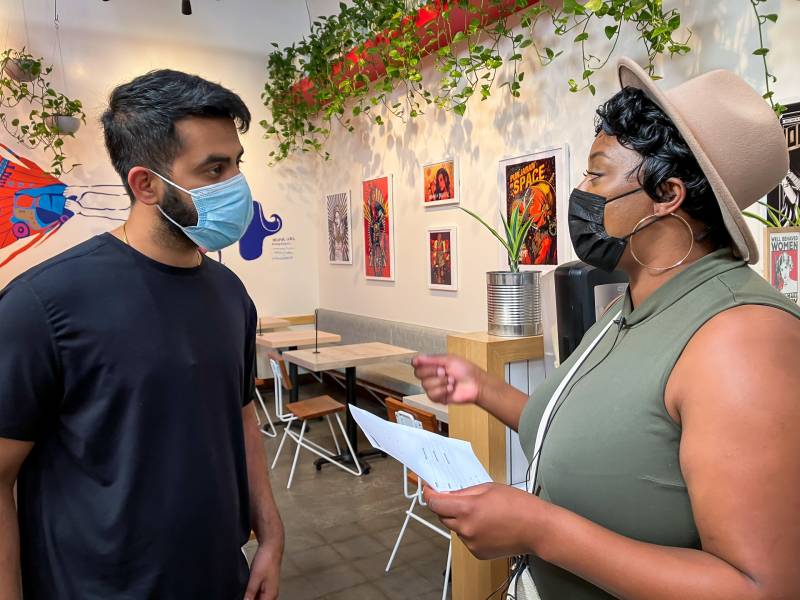 A Black woman with a green cutoff t-shirt and beige hat speaks through a black mask to a man in a blue surgical mask inside a bright space with tables and colorful art on the wall. 