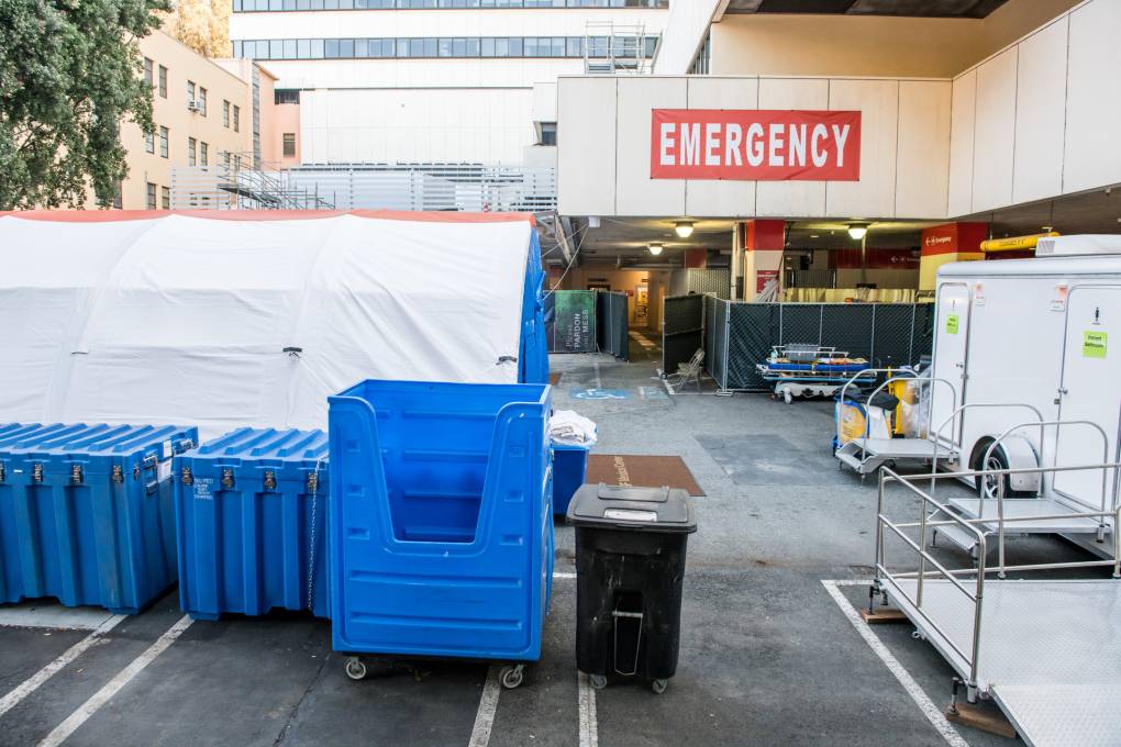 White tents lined with blue bins sit in front of the entrance to an emergency room.
